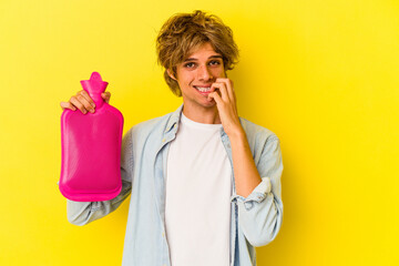Young caucasian man with makeup holding a hot bag of water isolated on yellow background  biting fingernails, nervous and very anxious.