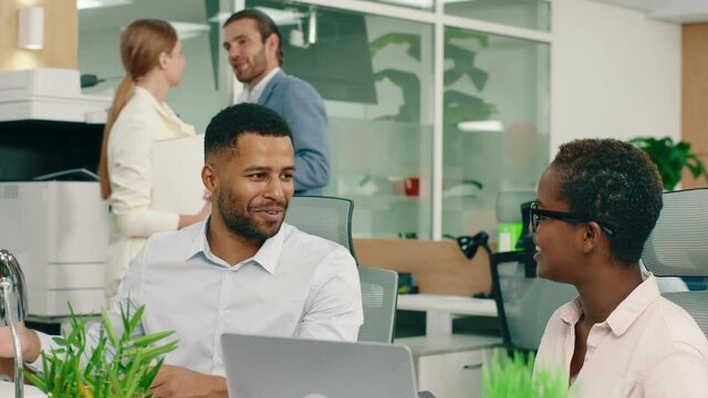 Two african workers are at the modern office and they are sitting at their desks smiling and agreeing with one another, in the back a man and woman are working at the printer