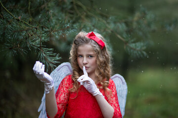 Weihnachtsengel im Märchen Wald, hält sich an der Tanne fest und lächelt. Das Engel hat blonde Locken, rotes Kleid an, rote Schleife. Sie hält Zeigerfinger vom Mund und bietet sie nicht verraten. 