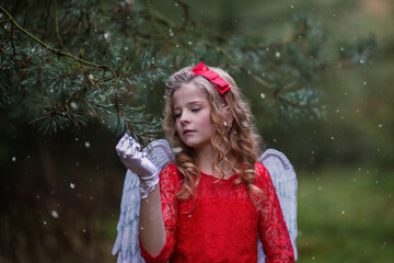 Weihnachtsengel im Märchen Wald, hält sich an der Tanne fest und lächelt. Das Engel hat blonde Locken, rotes Kleid an, Schneeflocken fallendes dem Himmel, sie hat eine Flocke gefangen, schaut sie an. 