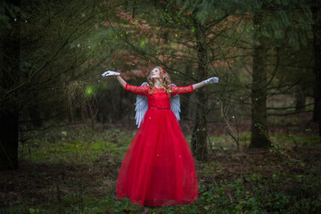 Weihnachtsengel in einem Märchen Wald. Es liegt kein Schnee. Typisch für Europa.  Das Mädchen hat ein rotes Kleid an, weiße Flügel und rote Schleife in der Haar. Sie tanzt, in der Hand Zauberlicht