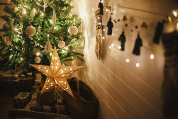 Stylish Christmas star and tree decorated with modern white baubles, boho ornaments and golden lights in atmospheric evening room. Festive scandinavian room at eve. Magic time. Space for text © sonyachny