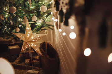 Stylish Christmas star, tree with white baubles, boho ornaments, golden lights and gifts in atmospheric evening room. Festive scandinavian room at eve. Magic time. Space for text. Merry Christmas! © sonyachny