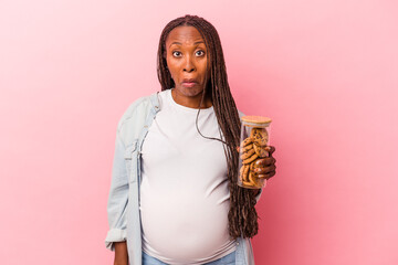 Young african american pregnant woman holding cookies isolated on pink background shrugs shoulders and open eyes confused.