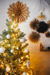 Stylish Christmas tree decorated with modern white baubles and golden lights on background of white wall with paper stars. Atmospheric festive scandinavian room. Christmas eve