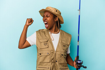 Young african american fisherwoman holding rod isolated on blue background raising fist after a...