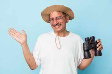 Middle aged indian fisherman holding rod isolated on blue background showing a copy space on a palm and holding another hand on waist.