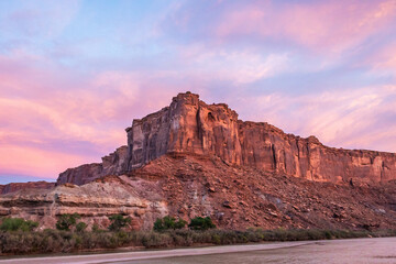 The brilliant colors of a sunrise reflecting on clouds over the top of a canyon wall with a river in the foreground