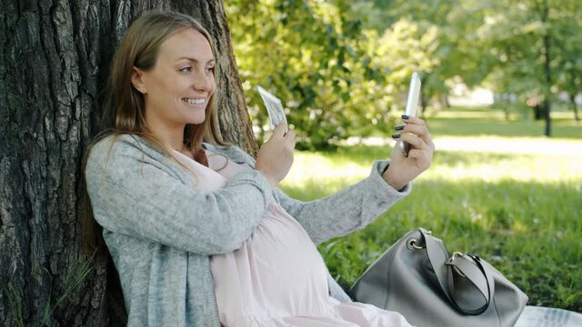 Emotional pregnant woman taking selfie with ultrasound picture of baby having fun outdoors in city park on summer day. Photo and pregnancy concept.