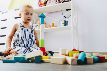 A boy with blonde hair in a jumpsuit plays children's educational wooden games in a bright room selective focus.