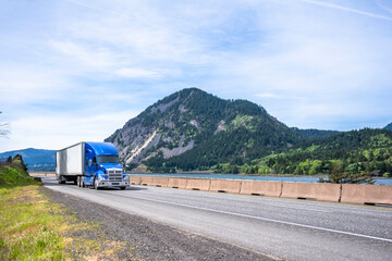 Professional industrial blue big rig semi truck transporting cargo in dry van semi trailer running on the road along river in Columbia Gorge area
