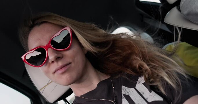 Young happy woman with heart shaped sunglasses making selfie video in car during road trip, goofing around. Customer generated video content.