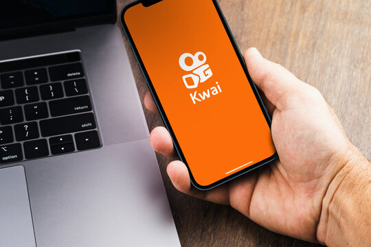 Male hand holding a smartphone with Kwai app on the screen with a computer beside it. Wooden Background. Rio de Janeiro, RJ, Brazil. August 2021.