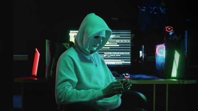 Portrait of a mysterious hacker in a hood, sitting at a table and looking at the camera after hacking a database in a dark room with animation of system codes in the background.
