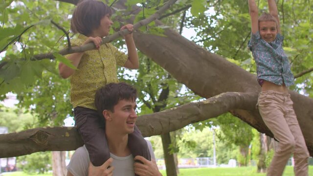 Cheerful father plays with children in the city park. The boy sits on his shoulders. A brother is hanging on a branch. A positive mood in an active game.
