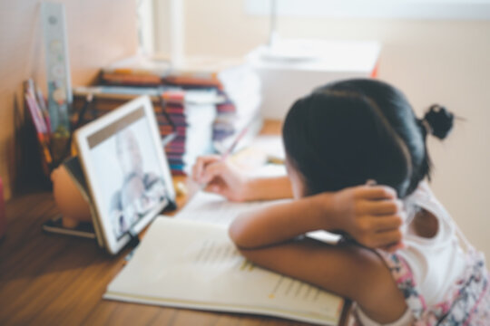 Blurred picture of Thai girl in online learning class at home during the COVID-19 situation. Student writing the book and learning form tablet during quarantine. Online education, home school concept.