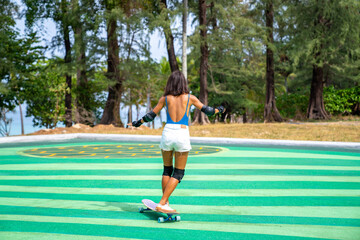 Attractive Asian woman with safety skateboarding knee pad skating at skateboard park by the beach. Happy female enjoy summer outdoor active lifestyle play extreme sport surf skate at public park.