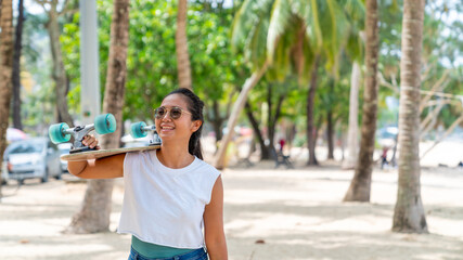 Portrait of Attractive smiling Asian woman holding skateboard walking on the beach in summer sunny day. Confidence female enjoy and having fun outdoor activity lifestyle and extreme sports surf skate