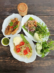View from the top of the dining table, Thai papaya salad, salmon salad, crispy and wooden pork belly, Bamboo container with sticky rice. Fresh vegetables and seafood sauce on wooden table. is a popula