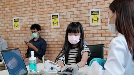 Asian female doctor with face mask checking the girl's health, measuring blood pressure, and use a stethoscope before the virus vaccination people were queuing in the back in the hospital clinic.