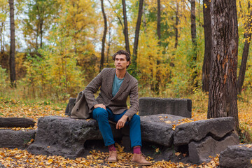 Tall handsome man dressed in a brown jacket posing with a bag on the rocks in autumn forest