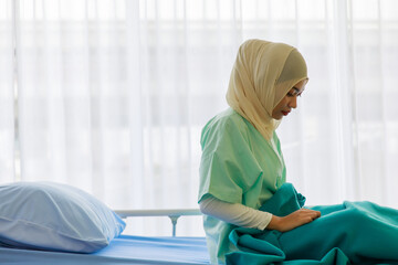 Portrait of sick young female Asian Muslim patient sitting on hospital bed wearing patient gown and cover with green blanket. Bright background