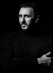 Black and white. Portrait of experienced adult man, writer in black turtleneck, sweater holding arms crossed at chest looking at camera over dark background