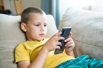 Young boy sitting on the sofa and playing or watching video in smartphone. Internet communication, modern technologies and people concept.