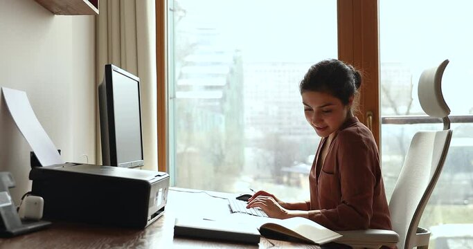 Side profile view young thoughtful Indian woman sit on comfy ergonomic chair looks in written notes, text on keyboard while studying working on pc thinks over task, doing remote work at workplace desk