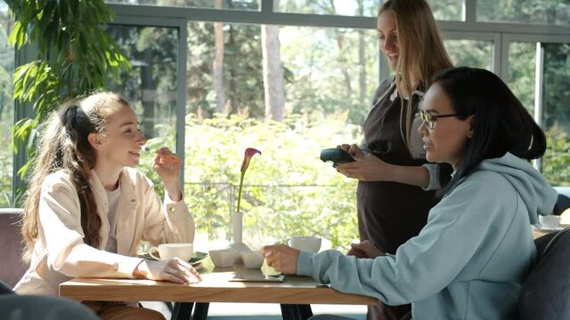 Young women friends are paying for lunch with credit card and talking to pregnant waitress in summer cafe. Modern technology and finance concept.