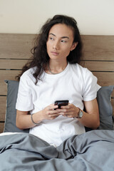 Handsome young man with long hair sitting in bed and texting friends in the morning