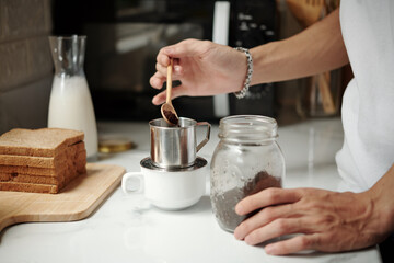 Hands of young man putting spoon of ground coffee in aluminum dripper on kitchen table