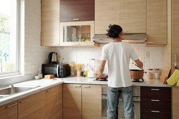 Young man in jeans and white shirt cooking dinner at kitchen counter, view from behind