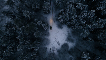 Aerial view of road and car in winter night forest. Forest work Establishing shots of a car in the winter landscape. Icy road. Snow covered nature. Spruce forest Latvia Winter background from above. 
