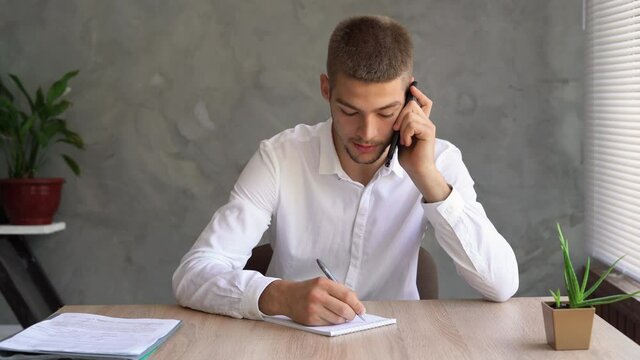 Caucasian guy business owner sitting at home in the office answers the phone call and makes a note with a pen in his notebook. The concept of phone sales, cold calls.