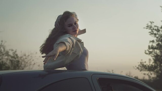 Young beautiful girl rides in the car with raised hands up, a woman in the sunroof of the car. Enjoying life at sunset in a car hatch. Hair in the wind in slow motion. Road trip adventure, wanderlust