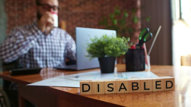 Wooden cubes with inscription disabled stand on office table against background of man in wheelchair, drinking coffee and working on laptop. Concept of possible work for people with disabilities