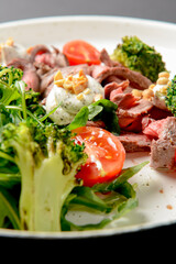 Healthy salad made from meat, cherry tomatoes, broccoli, goat cheese and fresh arugula served in a white plate.