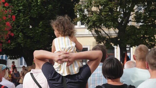 Father holds his child daughter on shoulders watching a city festival performance in a crowd of people spectators