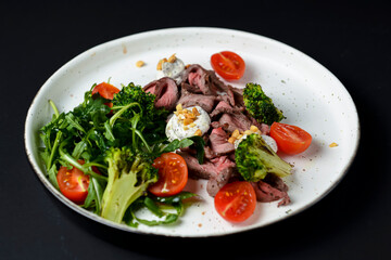Healthy salad made from meat, cherry tomatoes, broccoli, goat cheese and fresh arugula served in a white plate.