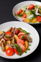 Two plates of vegetable salad made of tomatoes and fish slices, served in white plates over black background.
