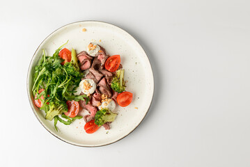Healthy salad made from meat, cherry tomatoes, broccoli, goat cheese and fresh arugula served in a white plate.