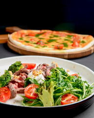 Healthy salad made from meat, cherry tomatoes, broccoli, goat cheese and fresh arugula served in a white plate.
