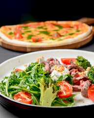 Healthy salad made from meat, cherry tomatoes, broccoli, goat cheese and fresh arugula served in a white plate.