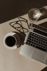 Aesthetic luxury bohemian minimalist home office workspace desk. Laptop computer, cup of coffee, glasses, crystal glass with sunlight shadows. Flat lay, top view work, business concept