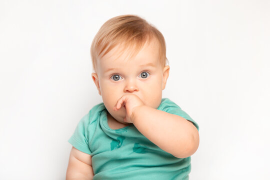 cute Caucasian one year old baby sucks thumb and scratches his first teeth. toddler boy teasing on white background in studio. baby care and child health concept