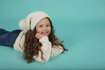 a beautiful happy brown-haired girl with curly hair in a white sweater and a white knitted hat lies on a blue background