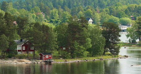 Many Red Swedish Wooden Sauna Logs Cabins Houses On Island Coast In Summer Cloudy Day