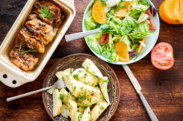 Boiled young potatoes with dill, fresh vegetable salad and baked chicken thighs on a dark wooden background.