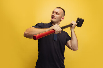 One young brutal man, firefighterposing with hammer over yellow studio background.